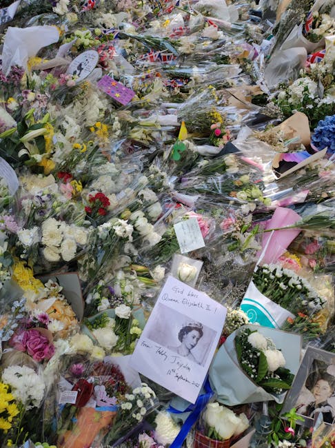 A large collection of floral tributes and bouquets arranged on the ground, primarily composed of white and pastel-colored flowers including roses, carnations, and chrysanthemums. The flowers are wrapp