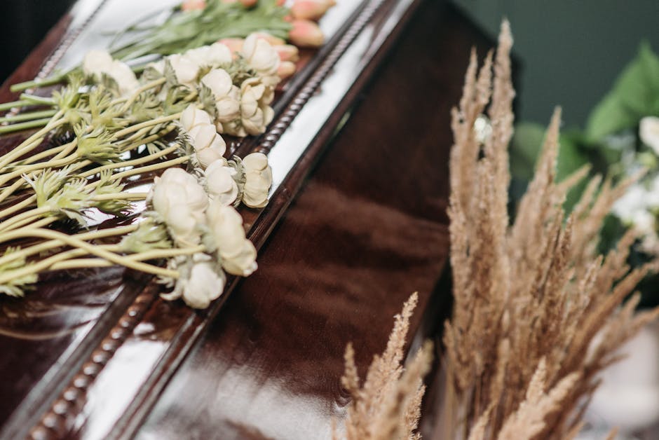 A close-up of a floral arrangement on a polished wooden surface, showcasing white ranunculus flowers with delicate, layered petals and long, slender green stems. The flowers are laid out in a linear f