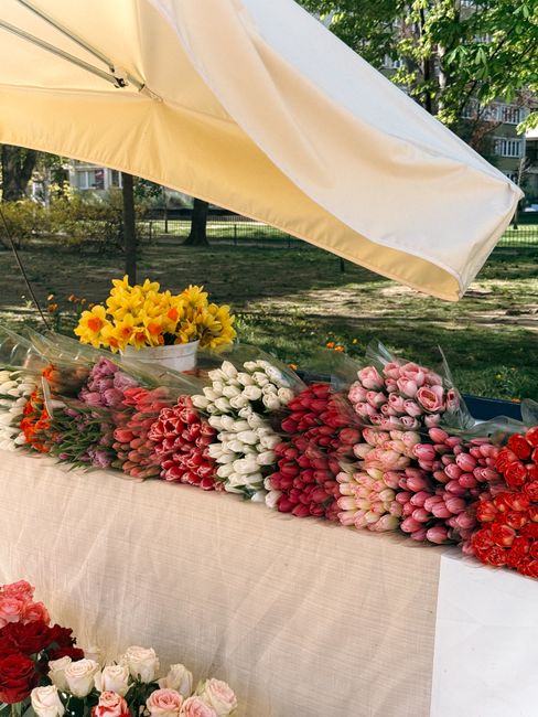 A vibrant display of fresh flower bouquets arranged on an outdoor market table beneath a large cream-colored umbrella. The flowers include tulips in shades of pink, white, and deep red, as well as clu