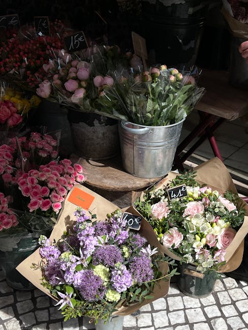 A street-side display of fresh flower bouquets arranged in various containers at a florist shop, showcasing a mix of pastel pink roses, lavender purple veronicas, and white flowers with green foliage.