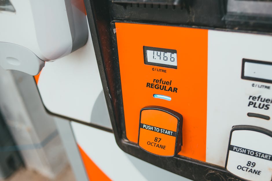 Close-up image of a fuel pump at a petrol station displaying a digital readout of 1466, with a bright orange and white color scheme. The pump features a black push-to-start button labeled '87 OCTANE' 